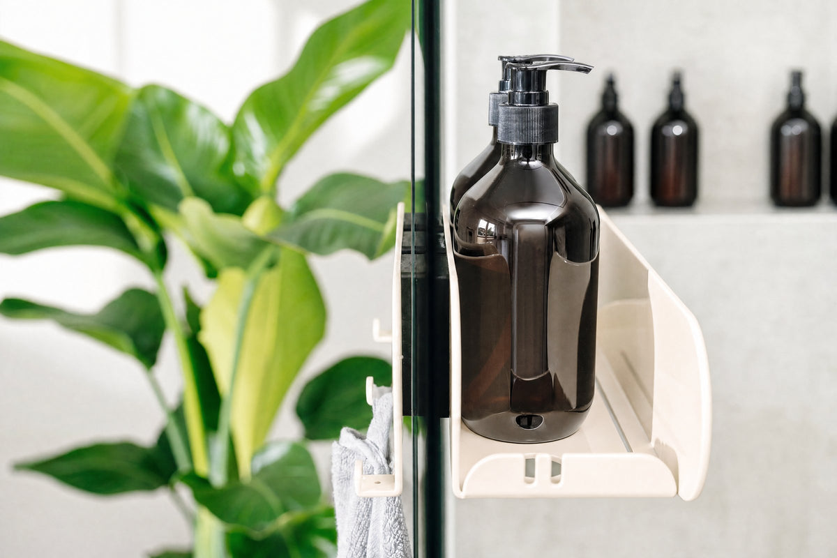 Duschablage ORBI Dock in Hellelfenbein hält eine Pumpflasche am Duschglas – Nahaufnahme